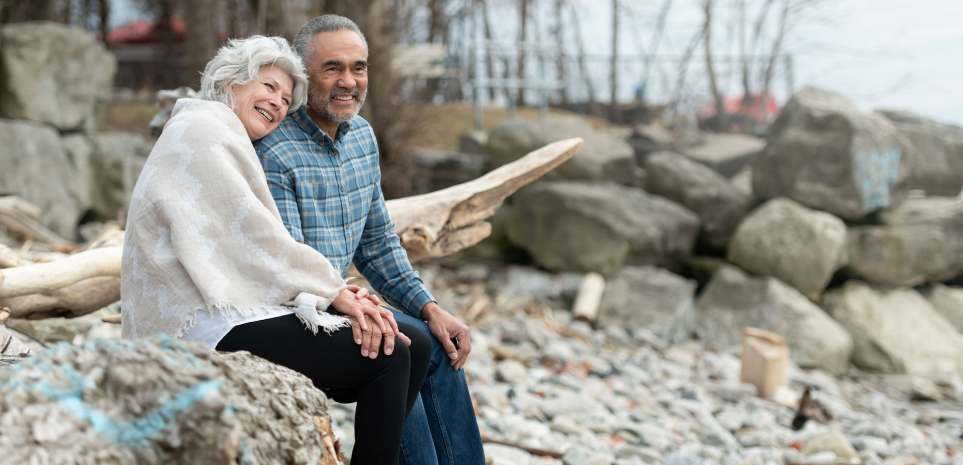 Aged couple shares smiling while sitting together on rocky beach.